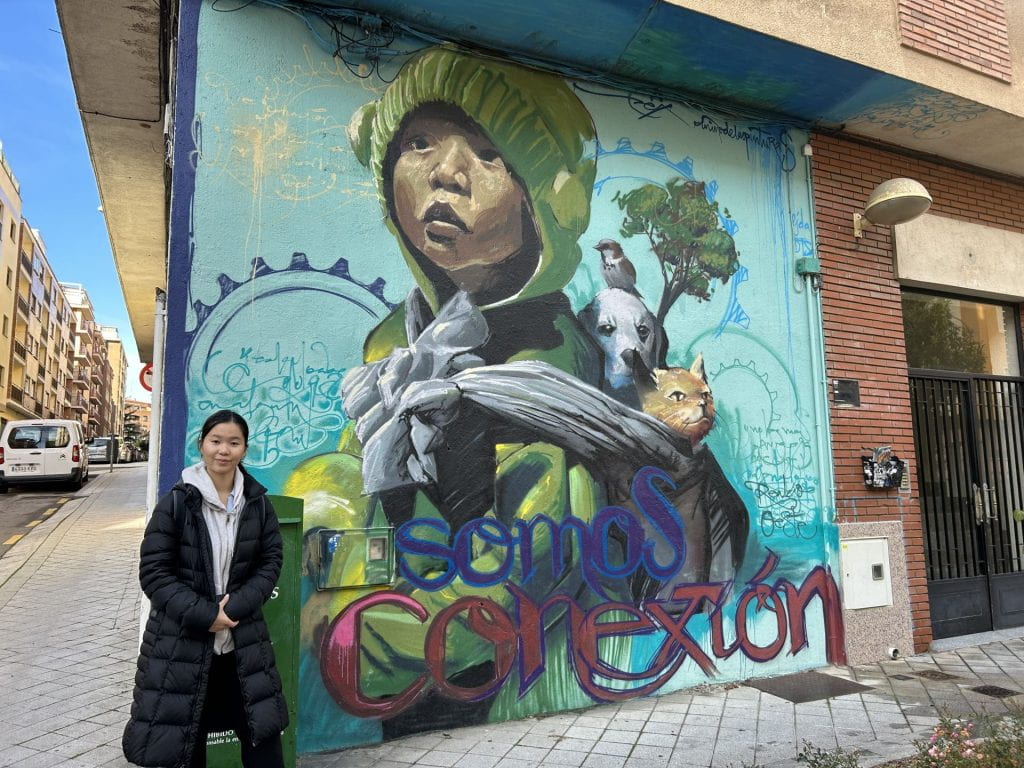 Woohyun standing in front of a mural which depicts a child carrying a dog, cat, and bird in a makeshift bag on their back. 