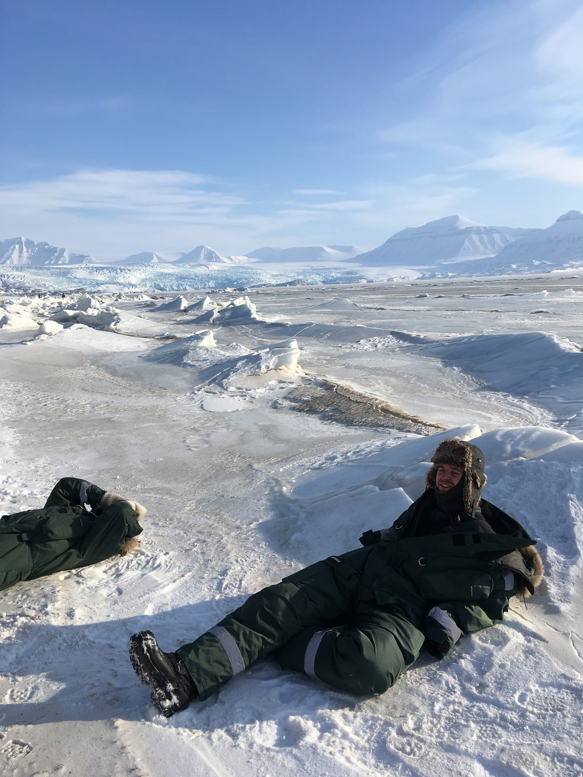 Two people bundled in winter clothing lounging on bed of snow and ice with blue sky in background